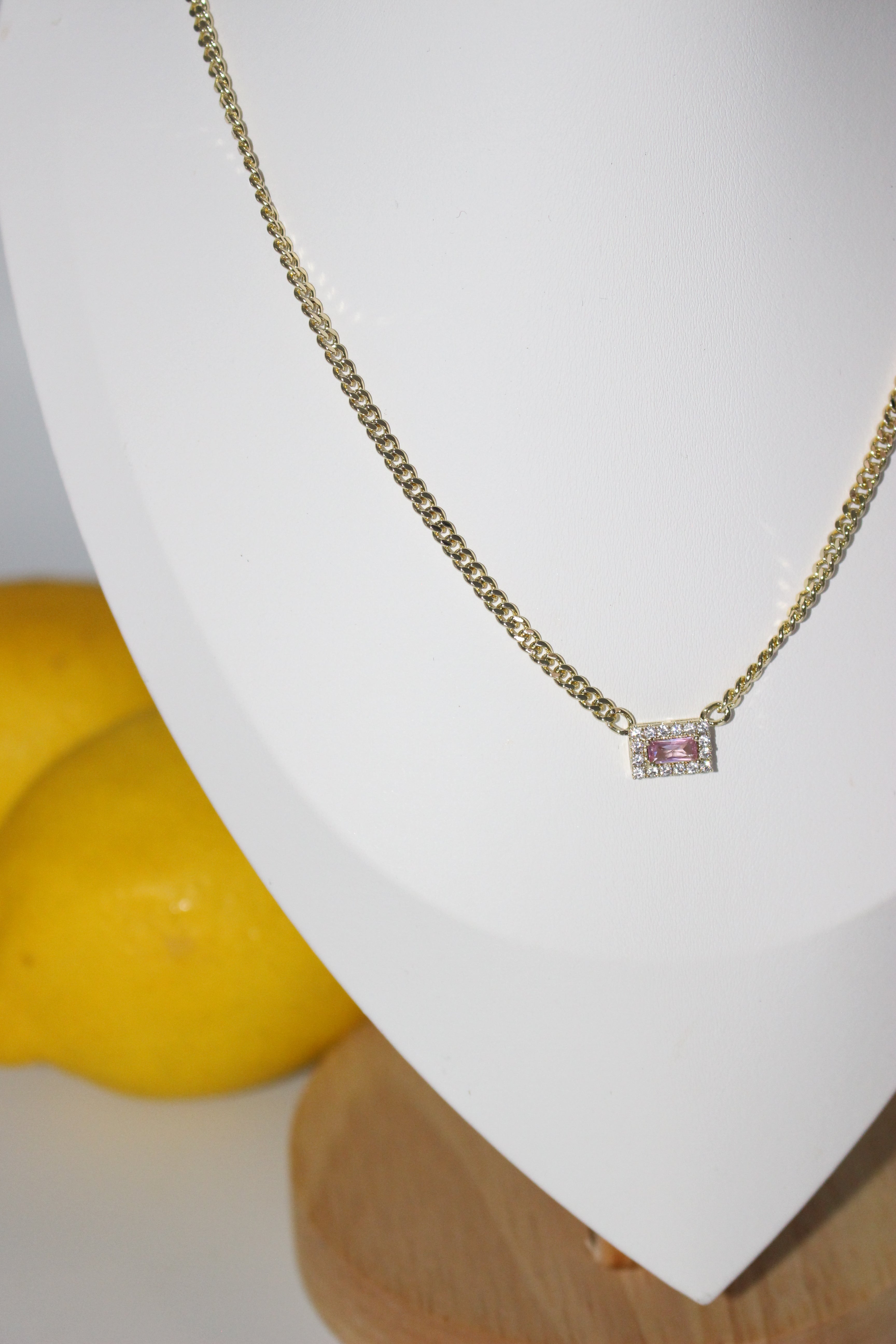 Gold necklace with a pink gemstone on a white cushion with a yellow fruit in the background