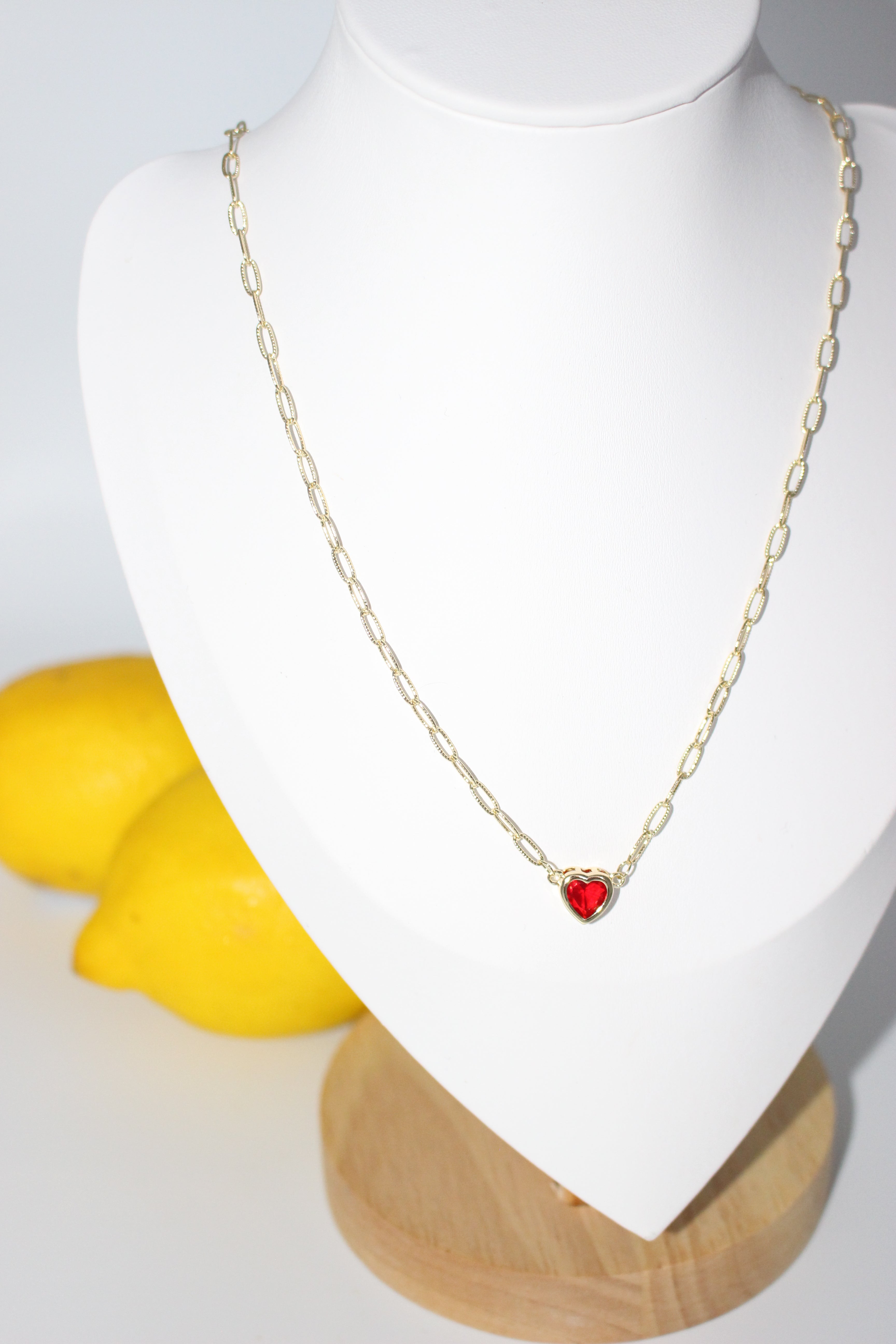 Gold necklace with a red pendant on a white stand with a yellow fruit in the background